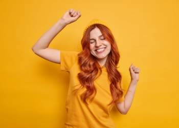 Studio shot of carefree redhead woman raises hand dances and smiles cheerfully has fun wears hat and casual t shirt isolated over vivid yellow background partying in good mood. Monochrome shot.