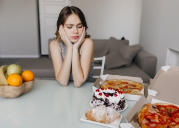 Sad curly woman looking at cake during diet. Blonde gorgeous female model posing with fruits and pizza.