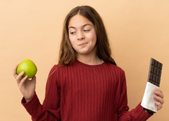 Little caucasian girl isolated on beige background having doubts while taking a chocolate tablet in one hand and an apple in the other